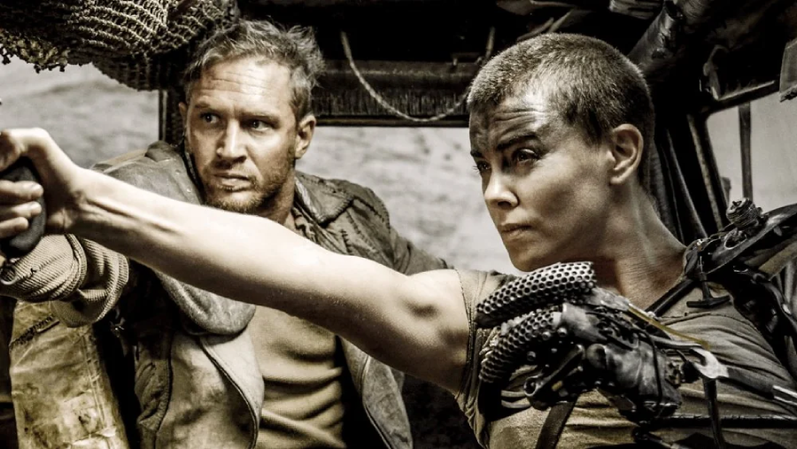 Two people in rugged, post-apocalyptic clothing sit inside a vehicle. The woman in front has a shaved head and is aiming a gun, while the man behind her looks alert and tense. Both appear determined and intense.