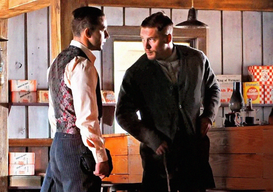 Two men in vintage clothing stand in a rustic, wooden-paneled room. One man faces the other while adjusting his coat, and vintage product boxes are visible on shelves in the background.