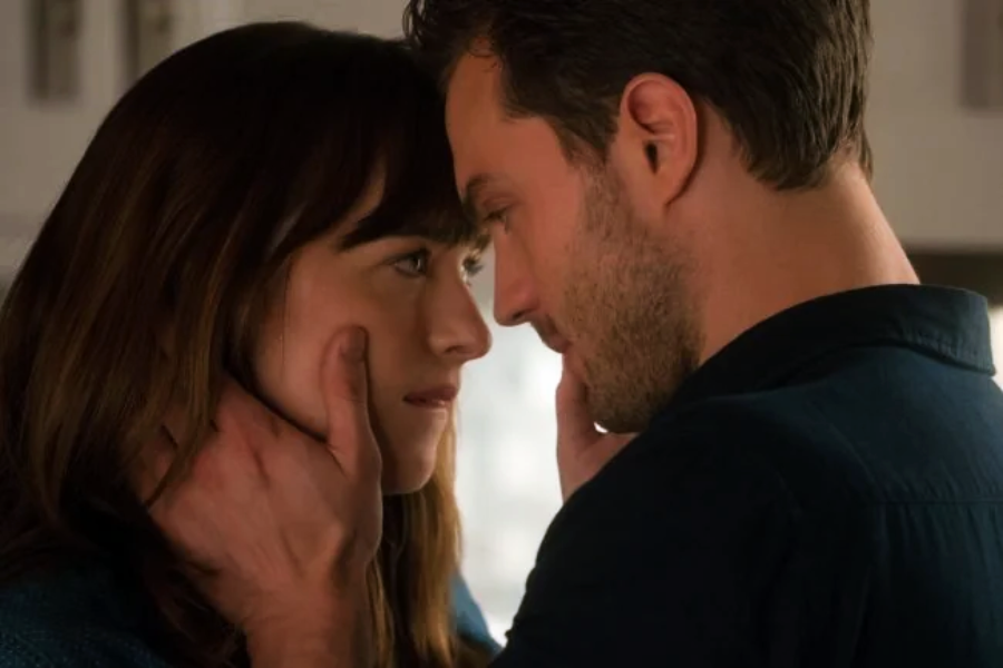 A man gently holds a woman's face as they stand close, gazing into each other's eyes in an intimate moment indoors.