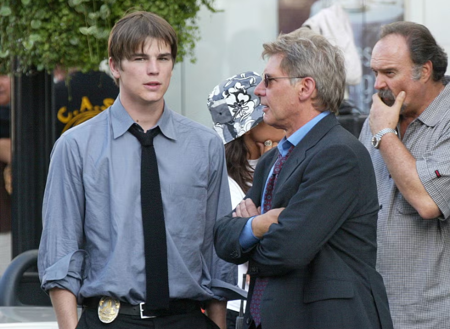 A young man in a police badge and tie stands next to an older man in a suit and sunglasses. They are outdoors, conversing, with two other people and greenery in the background.