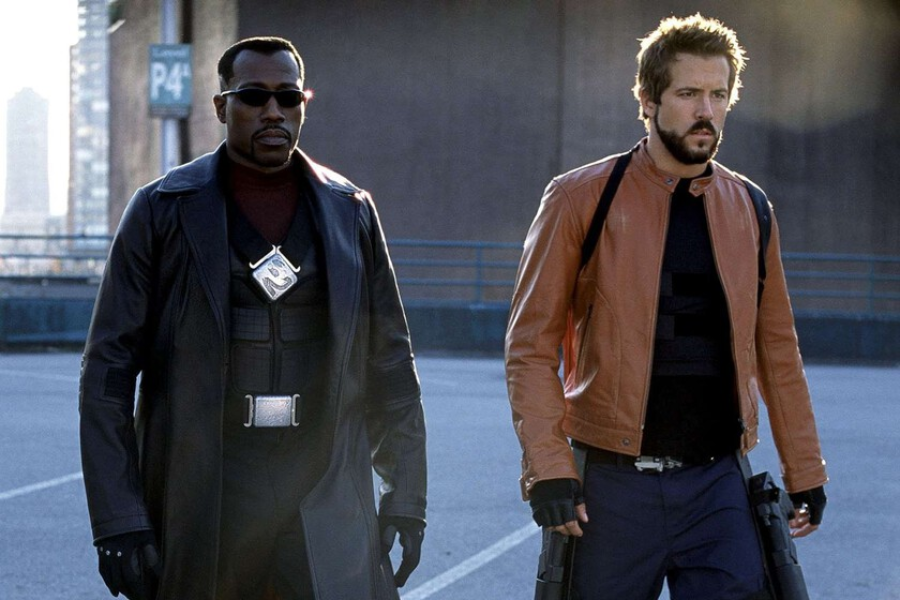 Two men in dark, futuristic outfits walk together in an urban setting. One wears sunglasses, a long black coat, and black gloves, while the other has a brown jacket, fingerless gloves, and a rugged appearance, both looking serious.