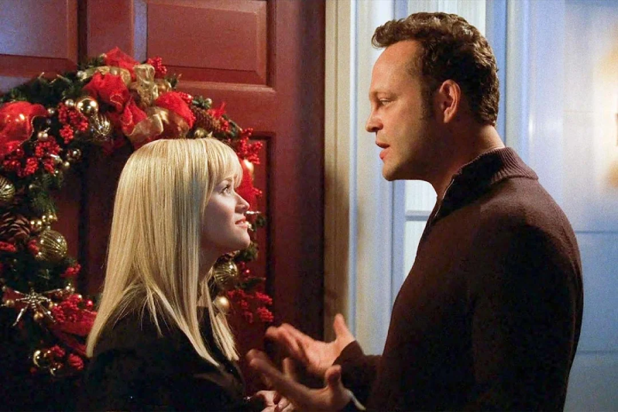 A woman with long blonde hair and a man with short brown hair stand talking near a red door decorated with a festive Christmas wreath. Both appear to be engaged in conversation, with holiday lights visible in the background.