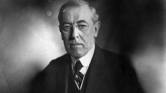 Black and white portrait of an older man in a suit and tie, looking directly at the camera with a serious expression. The background is blurred.