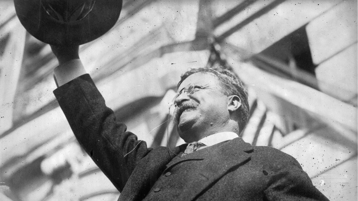 A man in a suit and glasses smiles broadly while raising a hat in the air, with a building in the background. The black-and-white photo is taken from a low angle.