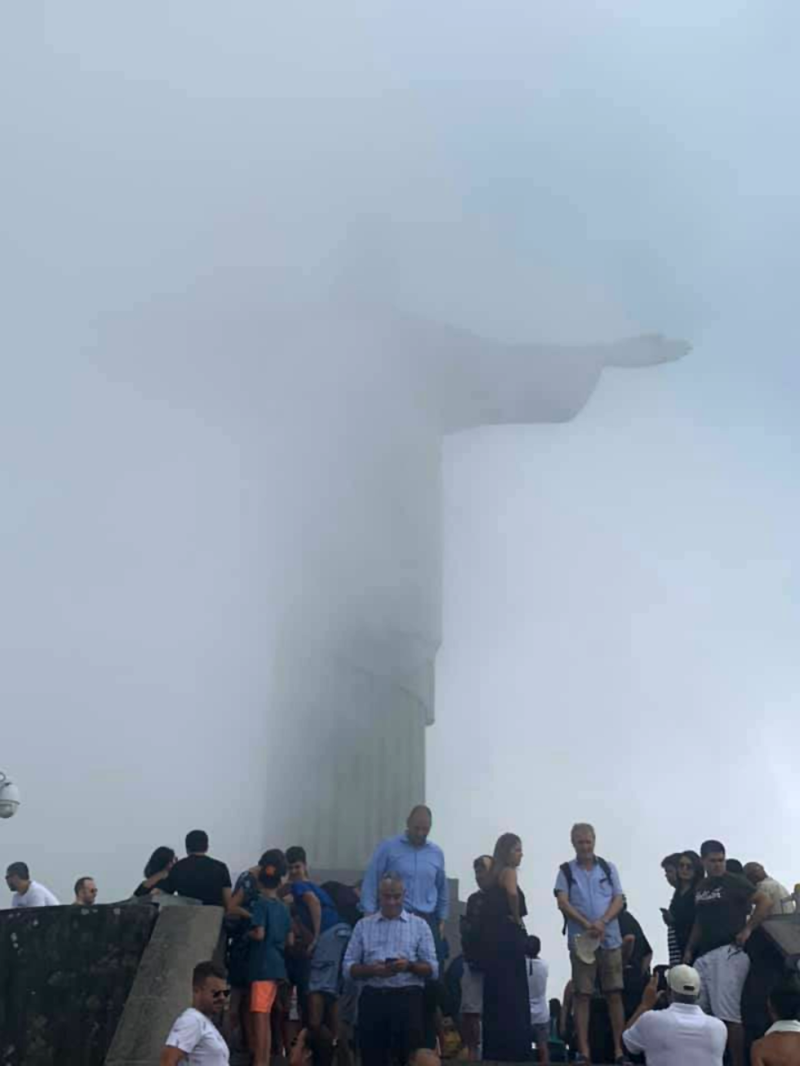 A crowd of tourists stands in front of the Christ the Redeemer statue, which is largely obscured by thick fog, rendering only the outline of the statue's arms and head faintly visible.