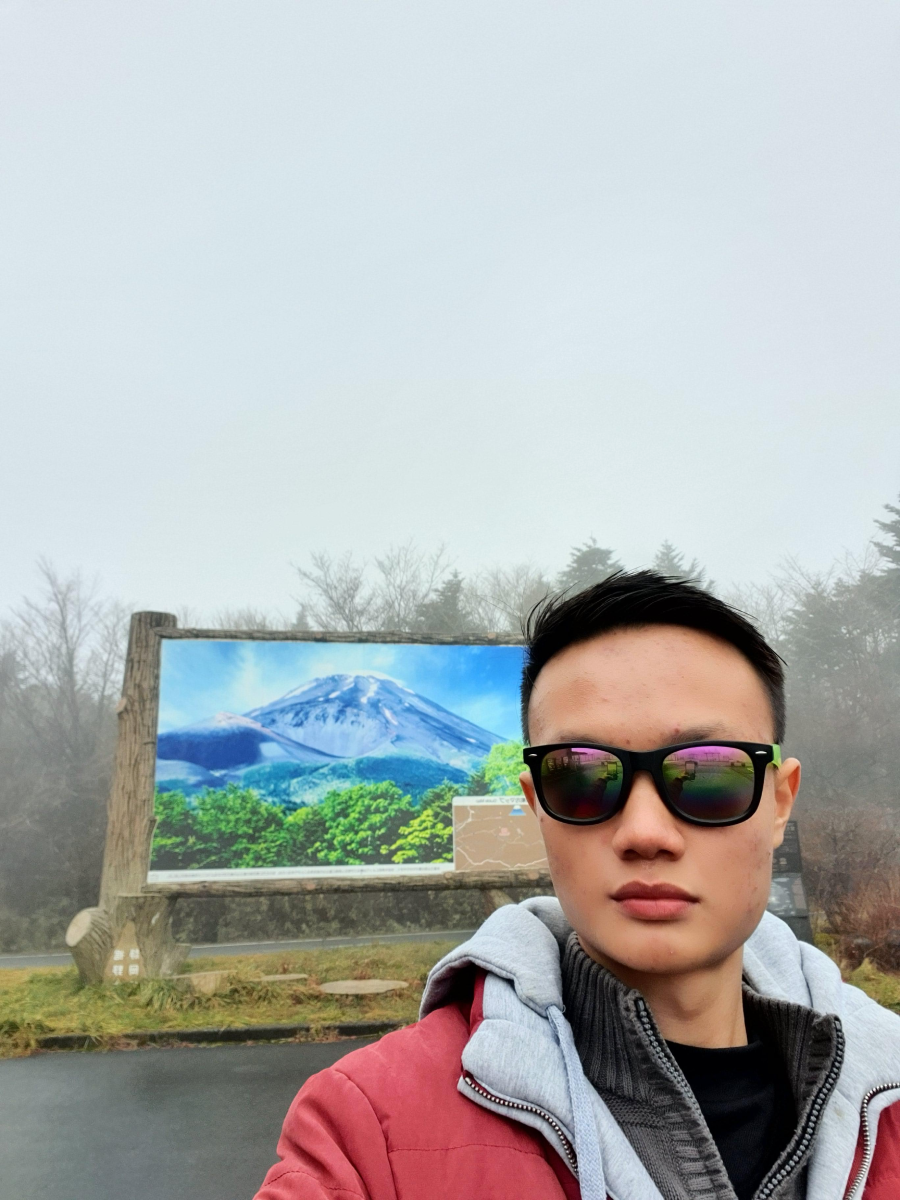 A person wearing sunglasses and a red jacket takes a selfie in front of a large sign featuring an image of Mount Fuji, with foggy trees and overcast weather in the background.