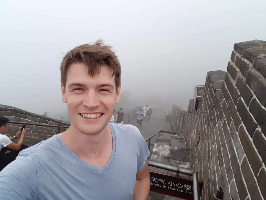 A young man smiles for a selfie on the Great Wall of China, with misty fog obscuring the background and a few tourists visible behind him.