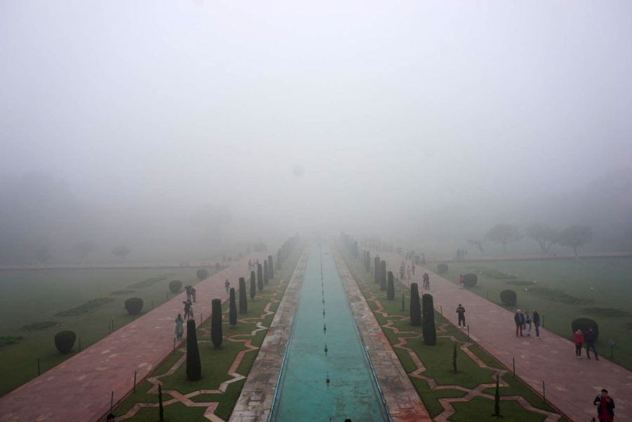 A long rectangular pool lined with small fountains and trees stretches into dense fog, with people walking along pathways on either side in a formal garden setting. Visibility is low due to the thick mist.