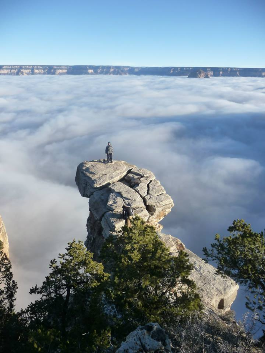 Two people stand on a rocky outcrop above a sea of clouds, with shrubs and trees in the foreground and distant cliffs visible on the horizon under a clear blue sky.