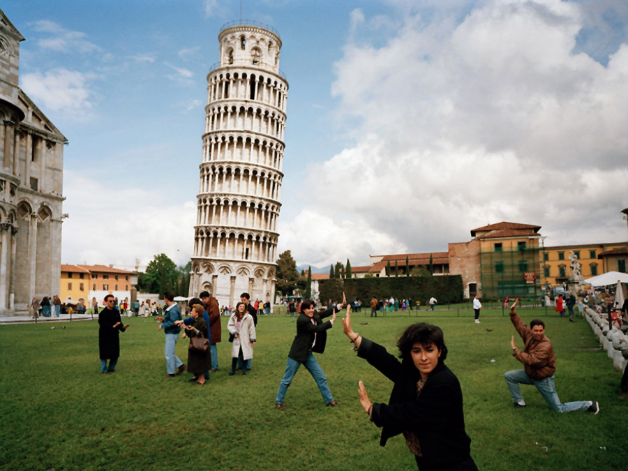 Tourists pose on a grassy lawn, playfully pretending to hold up the Leaning Tower of Pisa in the background on a partly cloudy day. The famous tower and nearby buildings are visible.