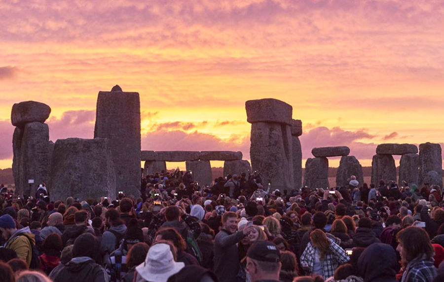 A large crowd gathers at Stonehenge during sunset, with people taking photos and the ancient stone structure silhouetted against a colorful sky.