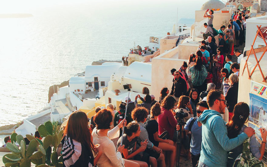 A large crowd of people gathers on a terrace overlooking white buildings and the sea at sunset, likely in Santorini, Greece. Some are taking photos, while others chat and enjoy the scenic ocean view.