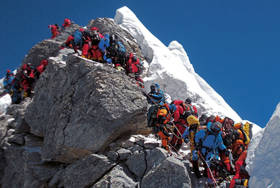 A large group of climbers in bright jackets and gear ascend a steep, rocky, and snowy mountain ridge in single file under a clear blue sky. Some use ropes and ladders to navigate the challenging terrain.