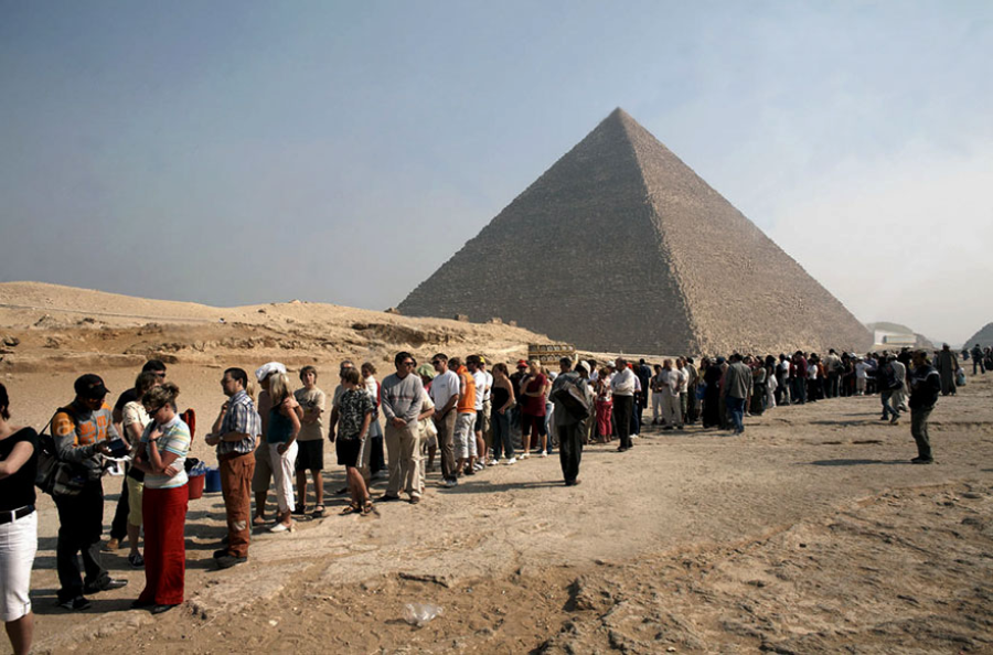 A long line of people waits near the Great Pyramid of Giza in Egypt, with the pyramid towering in the background under a hazy sky. The scene is set in a desert landscape.