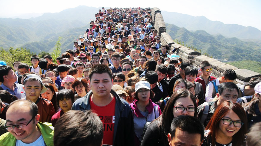 A large crowd of people closely packed together walks along a section of the Great Wall of China under a clear sky, with green mountains visible in the background.