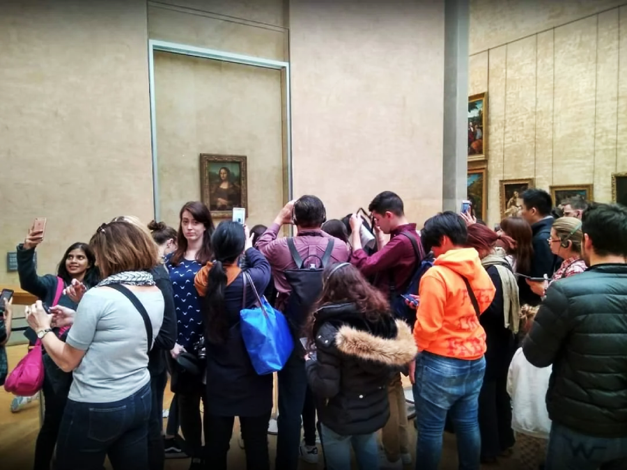 A group of people stand crowded in front of the Mona Lisa painting at a museum, many holding up their phones to take photos of the artwork.