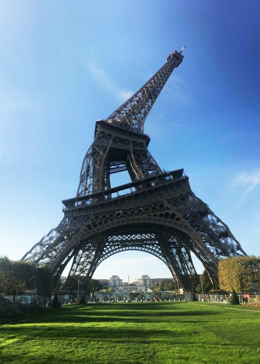 The Eiffel Tower appears bent and leaning to one side against a clear blue sky, with green grass and trees in the foreground.