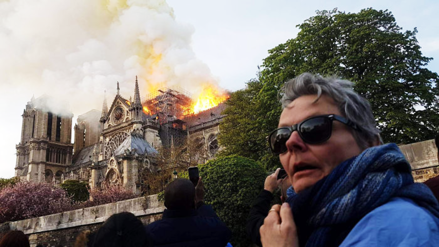 A person wearing sunglasses and a blue scarf stands in front of Notre-Dame Cathedral as it burns, with smoke and flames rising from the roof. Other people are taking photos, and trees frame the scene.