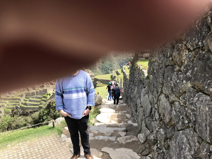 A person in a blue sweater stands on a stone path at Machu Picchu, with a large finger blocking the upper part of the image. Other tourists walk along a rocky path next to an old stone wall.