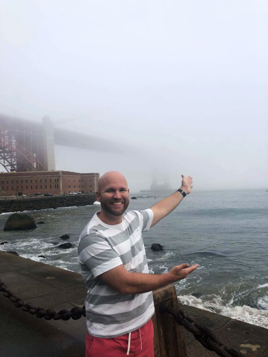 A man in a striped shirt and pink shorts smiles and gestures toward a foggy view of the Golden Gate Bridge, partially obscured by mist, near the shore.
