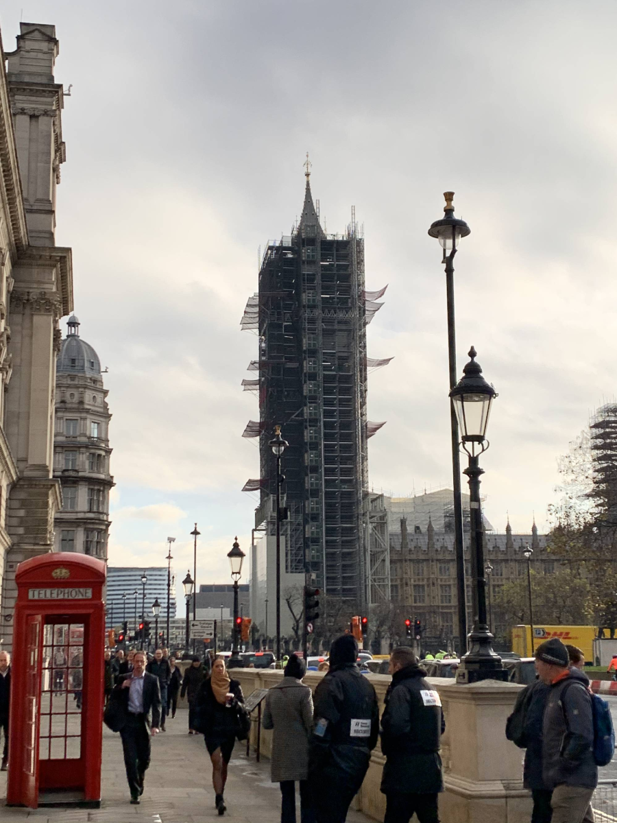 People walk along a city street in London near a classic red phone booth. In the background, Big Ben is covered in scaffolding for restoration, with cloudy skies overhead.