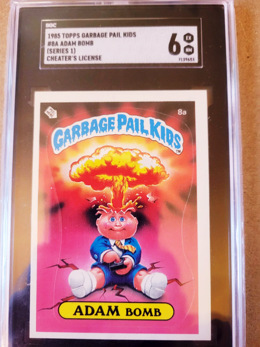 A graded 1985 Topps Garbage Pail Kids trading card (#8a ADAM BOMB) shows a cartoon child in a suit, sitting with an exploding mushroom cloud coming out of his head. The card is encased in a plastic holder.