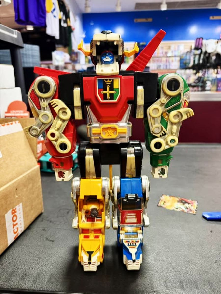 A vintage Voltron robot action figure stands on a store counter, assembled from colorful lion components, with shelves of merchandise visible in the background.