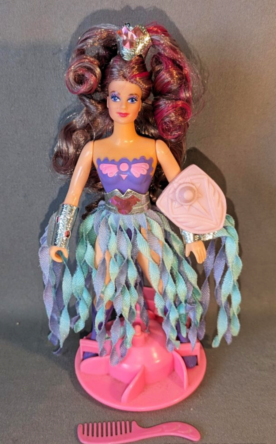 A Barbie doll with long curly brown and purple hair, wearing a purple and teal fantasy outfit with silver arm cuffs and a pink shield. She stands on a pink base, with a pink comb in front of her.