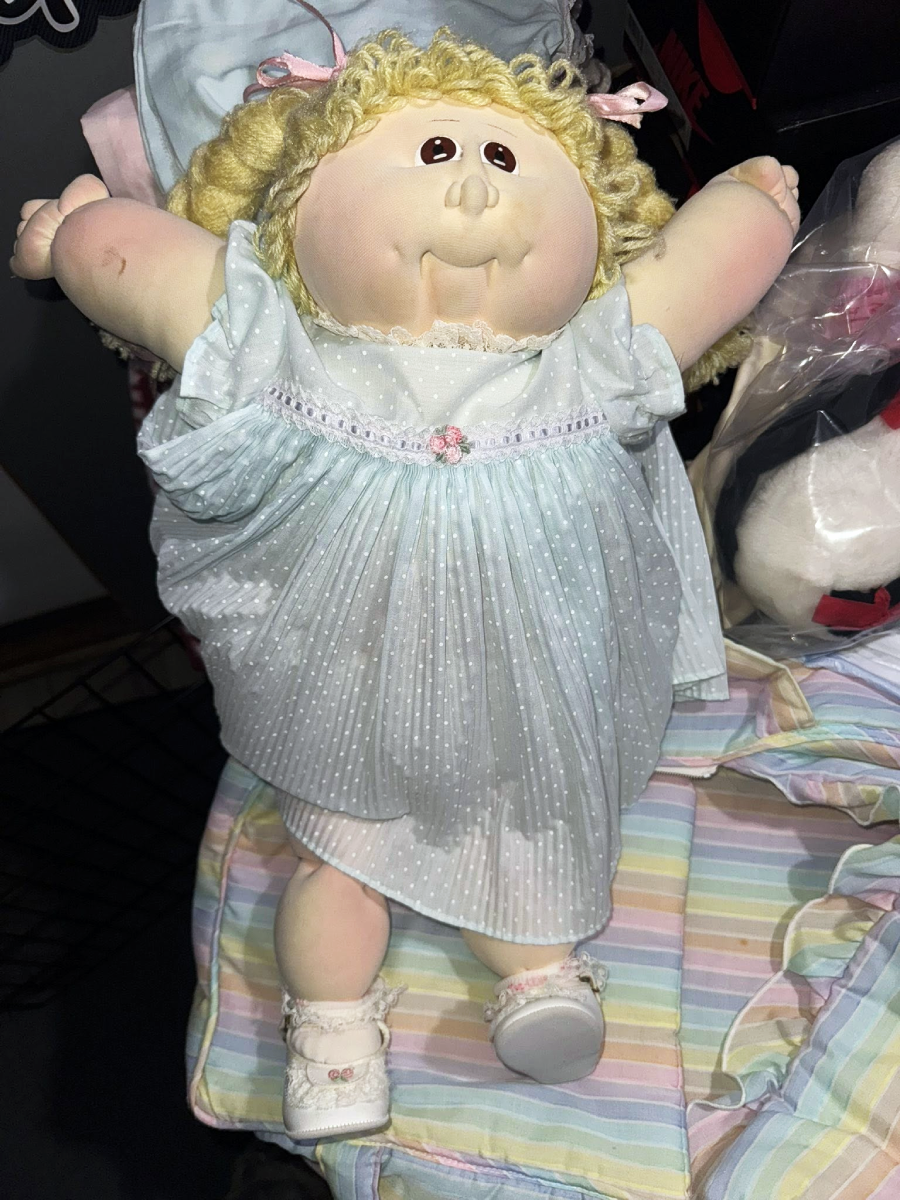 A Cabbage Patch Kids doll with curly blonde hair, wearing a pale blue dress with white polka dots and white shoes, is lying on a pastel-striped blanket with its arms raised.