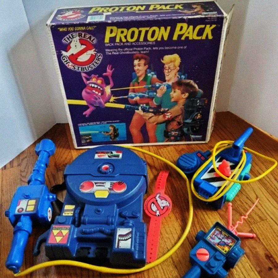 A vintage "The Real Ghostbusters" Proton Pack toy set is displayed on a wooden floor, with the box in the background. The set includes a blue proton pack, a blaster, a red wrist device, and connecting yellow tubes.