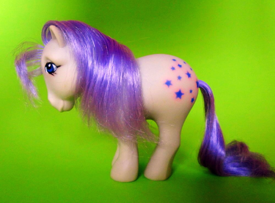 A toy pony with light beige body, purple mane and tail, and blue star markings on its side stands against a bright green background.