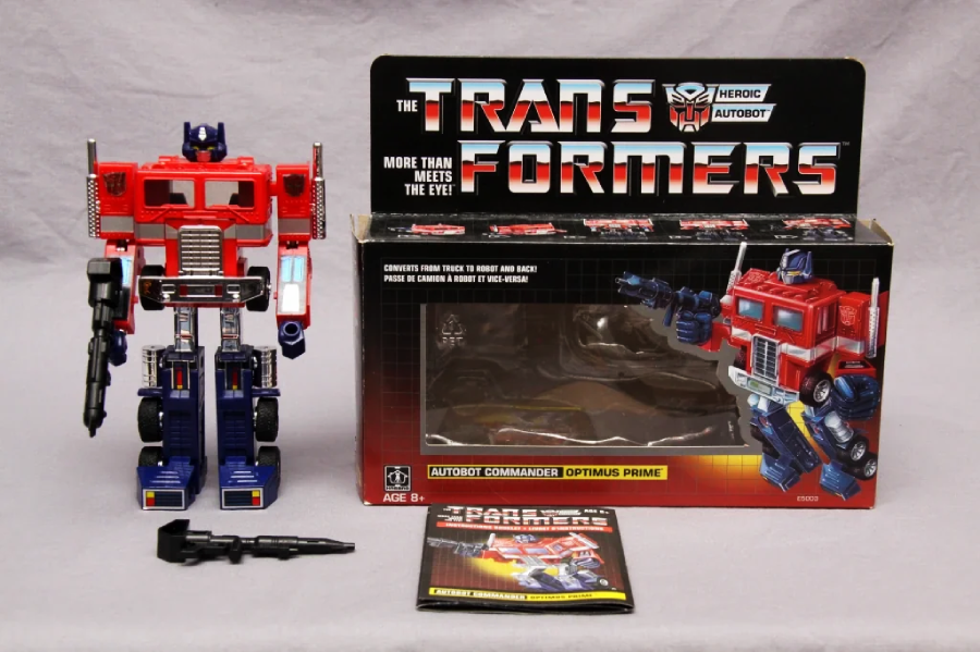 A vintage Transformers Optimus Prime action figure stands next to its open box and instruction manual. The box features Transformers branding, character art, and “Autobot Commander Optimus Prime” text. Accessories are also included.