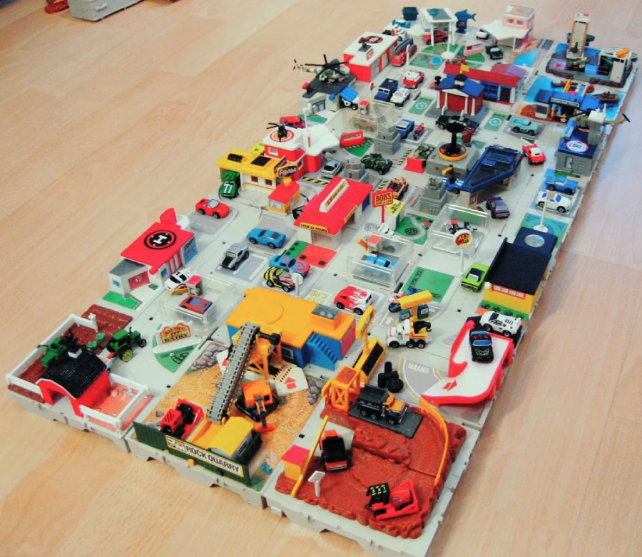 A colorful toy city playset with various miniature buildings, vehicles, and helicopters arranged on a board, set on a wooden floor. The scene includes cars, fire trucks, and small shops.