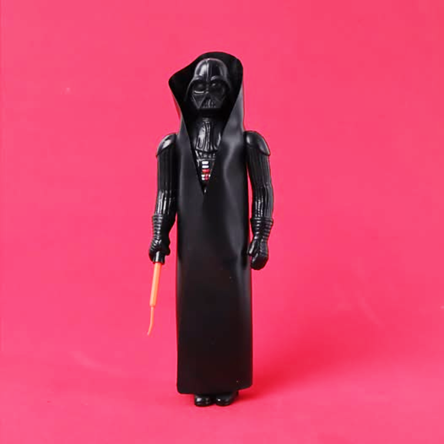 A Darth Vader action figure with a black cape and helmet, holding a small red lightsaber, stands against a solid pink background.