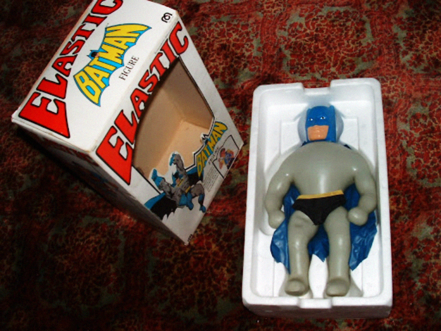 A vintage Elastic Batman figure lies in a white plastic tray next to its open box on a patterned red carpet. The box shows an illustration of Batman and bold text reading "Elastic Batman Figure.