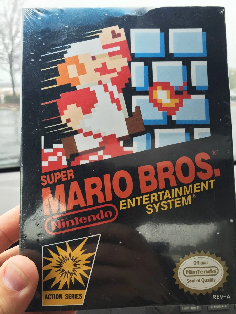A hand holds a sealed NES Super Mario Bros. video game box featuring pixel art of Mario breaking bricks, with the Nintendo and Action Series logos visible.