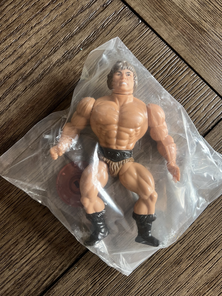 An action figure of a muscular man with black boots and a loincloth, packaged in a clear plastic bag, lies on a wooden surface.