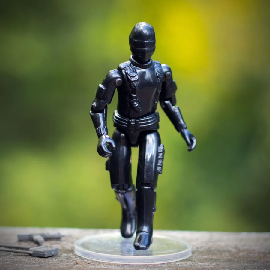 A black action figure resembling a robot or futuristic soldier stands on a clear round base outdoors, with a blurred green background. Small gray accessories lie on the surface nearby.