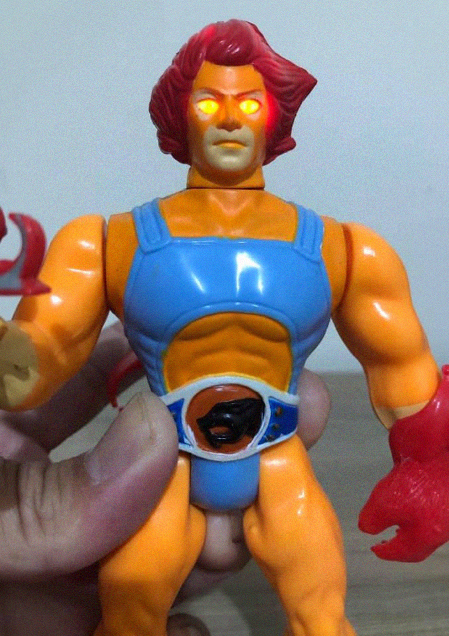 A hand holds an action figure with orange skin, red hair, glowing eyes, and a blue outfit. The figure has a red claw on its right hand and an emblem on its belt.