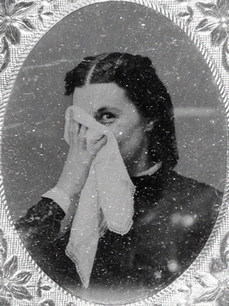 A vintage black-and-white portrait of a woman holding a handkerchief to her face, partially covering her mouth and nose, with a slight smile. The photo is framed with an ornate, floral border.