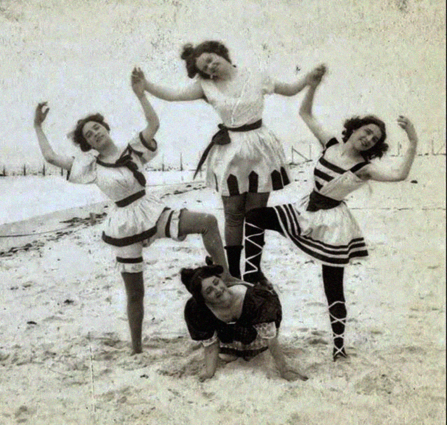 Four women in vintage bathing costumes pose playfully on a sandy beach, forming a human pyramid with three standing and one crouched at the base, all smiling and holding hands.