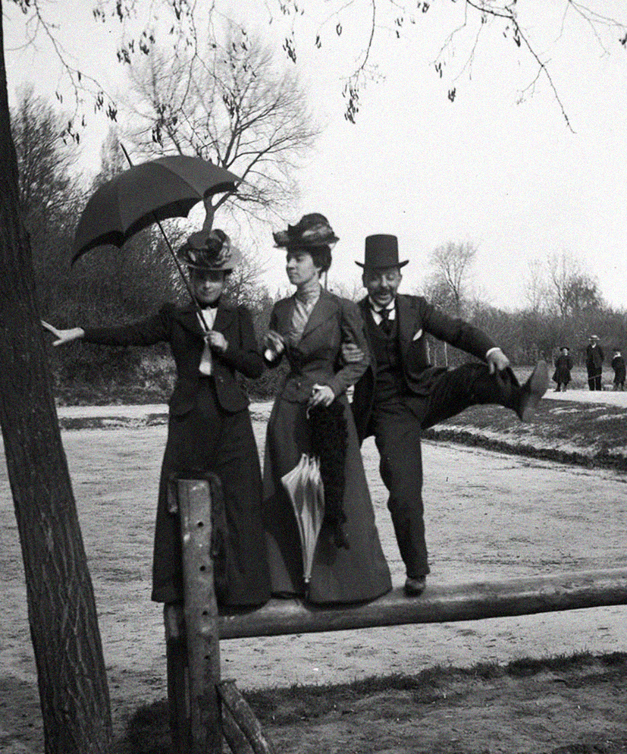 Three people in vintage clothing balance on a wooden fence. One woman holds an umbrella, another holds a closed parasol, and a man in a top hat lifts his leg playfully. Leafless trees and people are visible in the background.