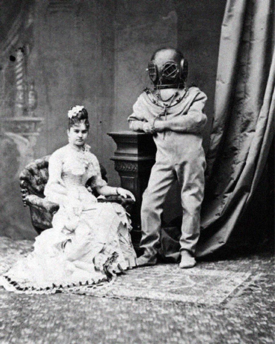 A vintage black-and-white photo of a seated woman in a Victorian dress beside a standing person wearing an old-fashioned deep-sea diving suit and helmet, posing indoors with draped curtains in the background.