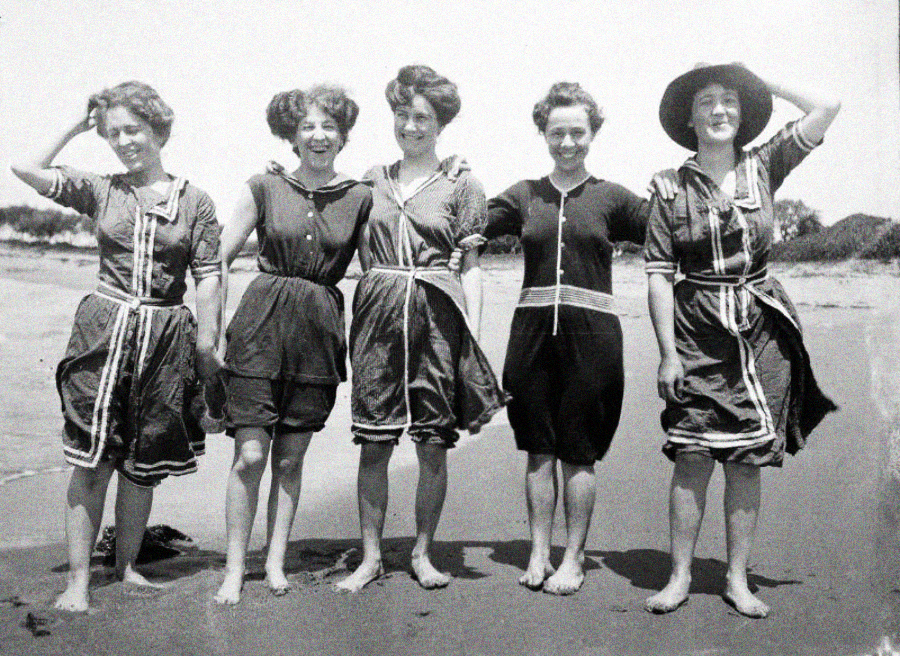 Five women stand barefoot on a beach, smiling at the camera. They wear vintage, modest bathing suits with short sleeves and knee-length shorts. The background shows sand, water, and distant trees under a bright sky.