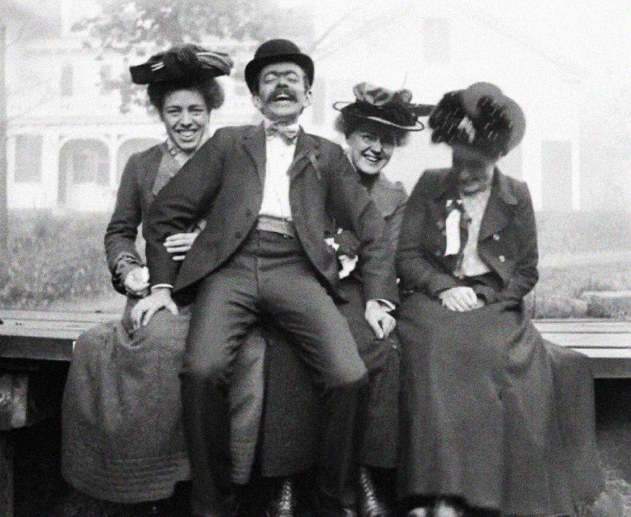 Four people in early 1900s attire, three women and one man, sit closely together on an outdoor bench, smiling and laughing. The women wear large hats, and a house is visible in the blurry background.