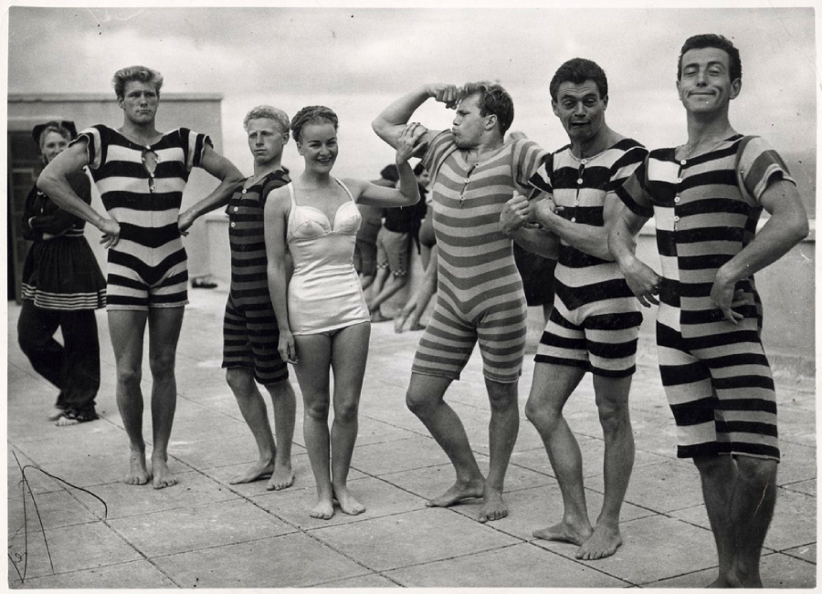 A group of men in striped vintage swimsuits pose playfully with a woman in a white swimsuit on a beach promenade, with more people and cloudy skies in the background.