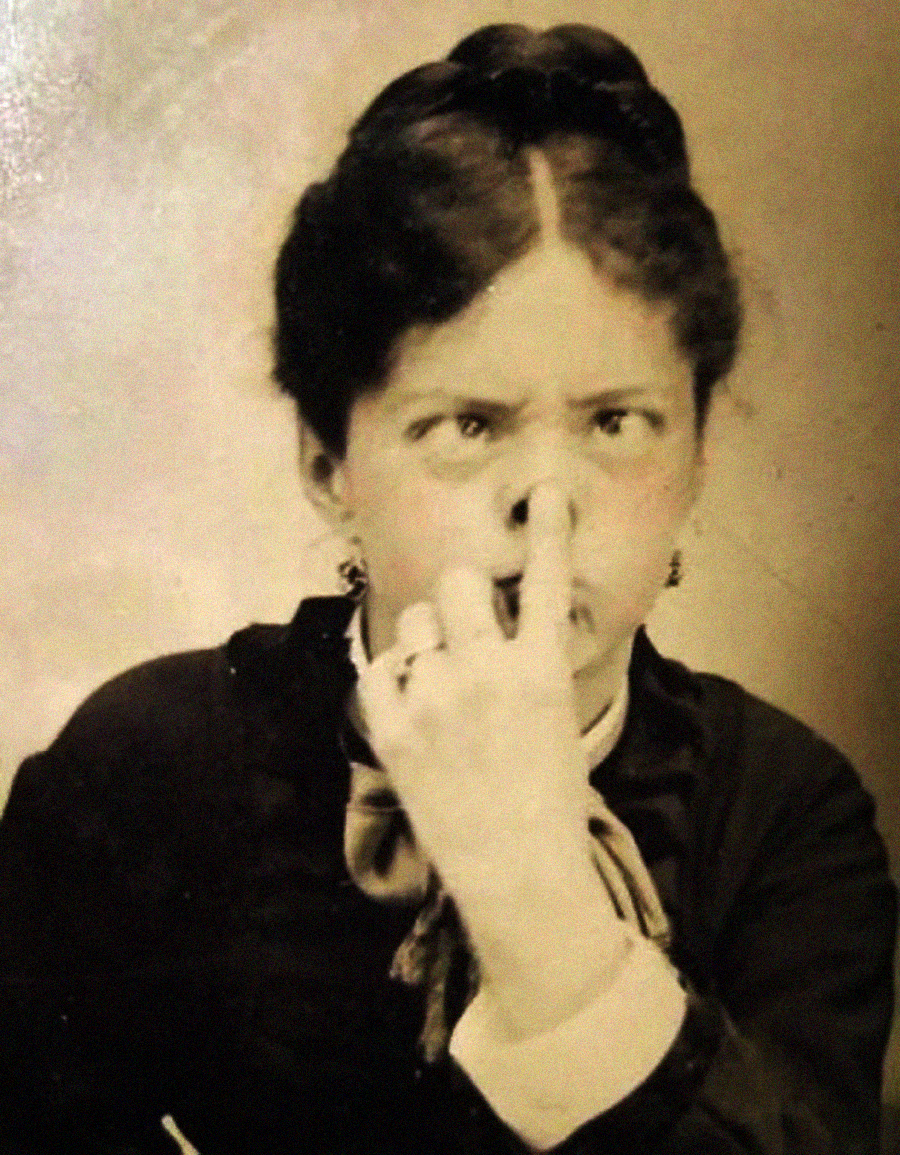 A vintage sepia photo of a woman with dark hair, wearing a dark dress, holding her nose with her fingers and making a playful, silly face at the camera.