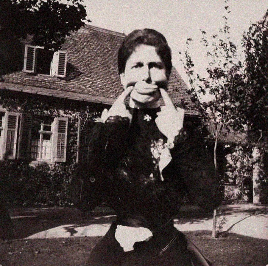 A black-and-white photo of a woman standing outside in front of a house, wearing a dark dress and pulling the sides of her mouth into a wide smile with her fingers. Trees and a garden are visible in the background.