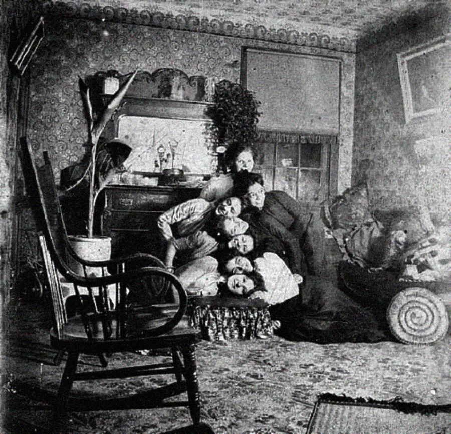 A black-and-white photo shows several people piled on top of each other in a cozy, cluttered vintage living room with patterned wallpaper and assorted furniture.