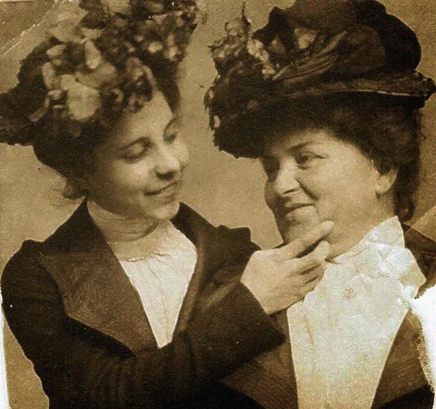 A sepia-toned vintage photo shows two women wearing hats decorated with flowers. One woman gently touches the other's chin as they smile warmly at each other, dressed in high-collared blouses and jackets.